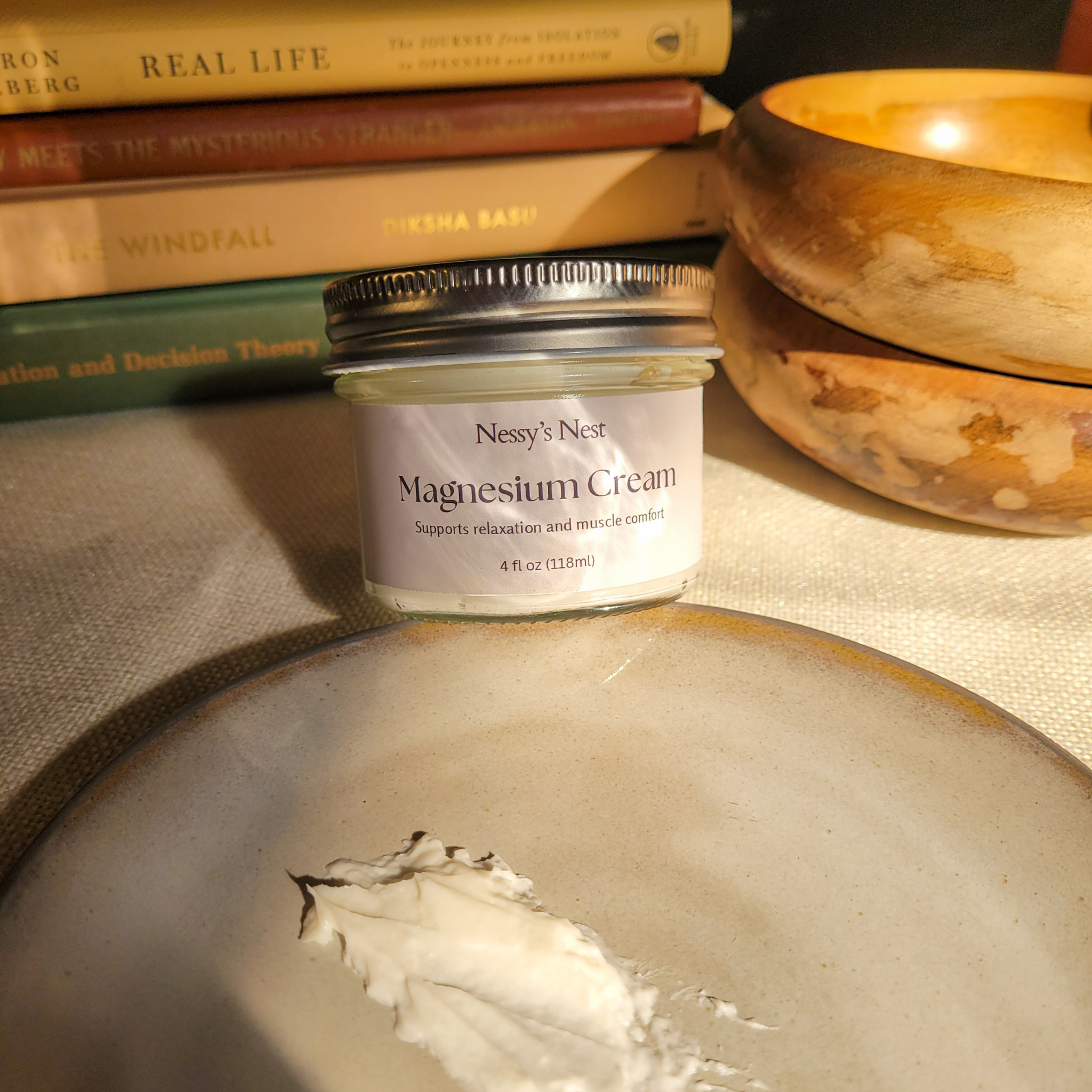 Jar of Nessy's Nest Magnesium Cream on a textured surface with books and wooden bowls in the background.