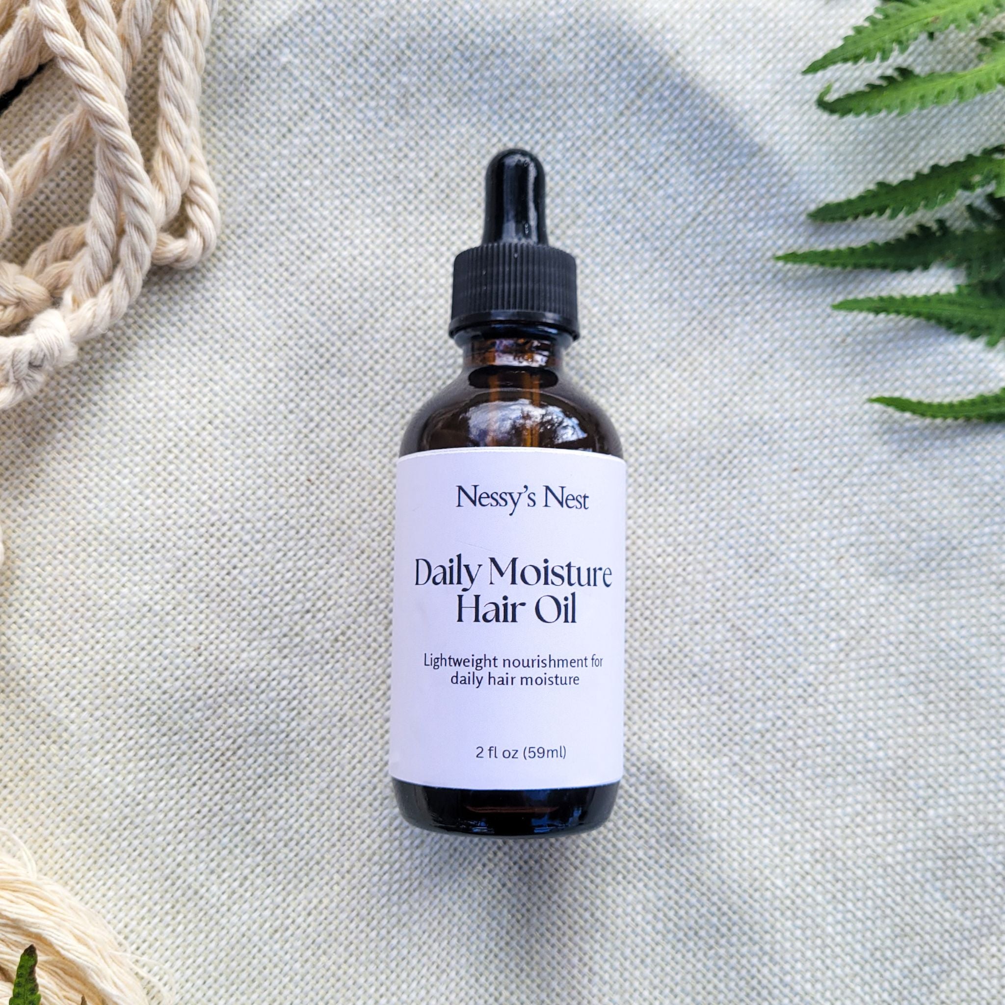 Bottle of Nessy's Nest Daily Moisture Hair Oil on a textured light background with green leaves.