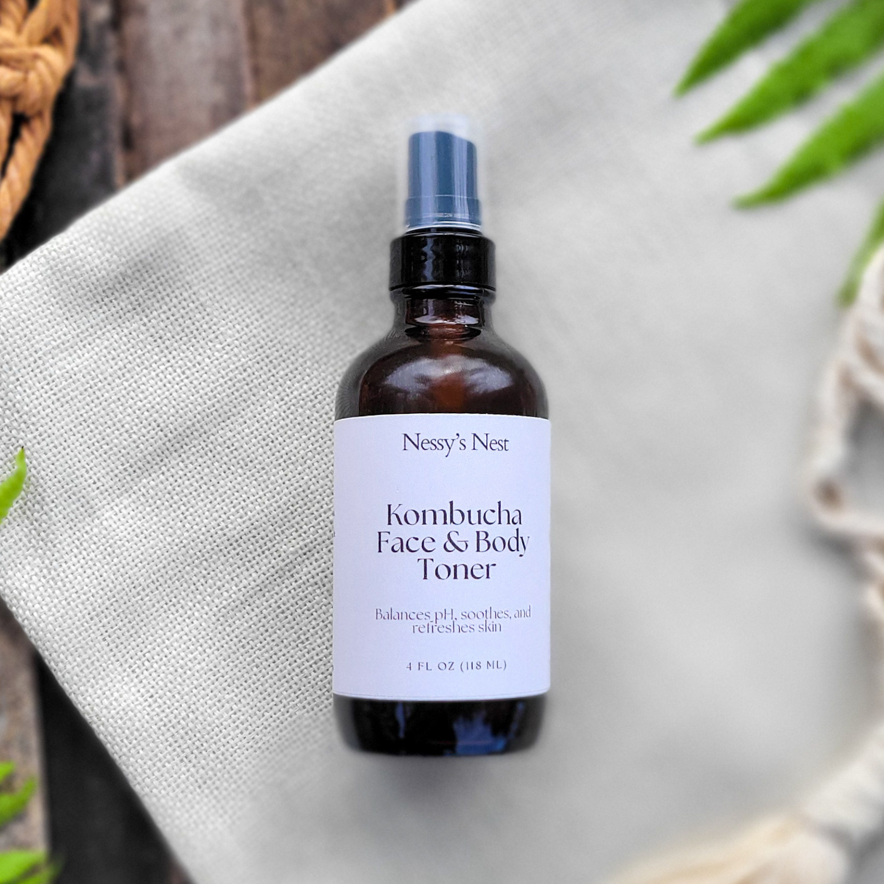 Nessy's Nest Kombucha Face & Body Toner bottle on a textured surface with greenery.